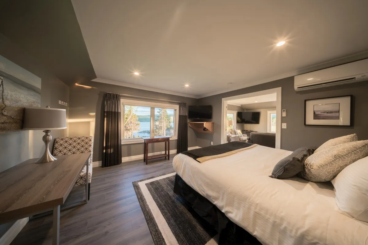 A bedroom with a king bed, a desk and lamp beside a window looking onto the water, and a doorway into the sitting room.