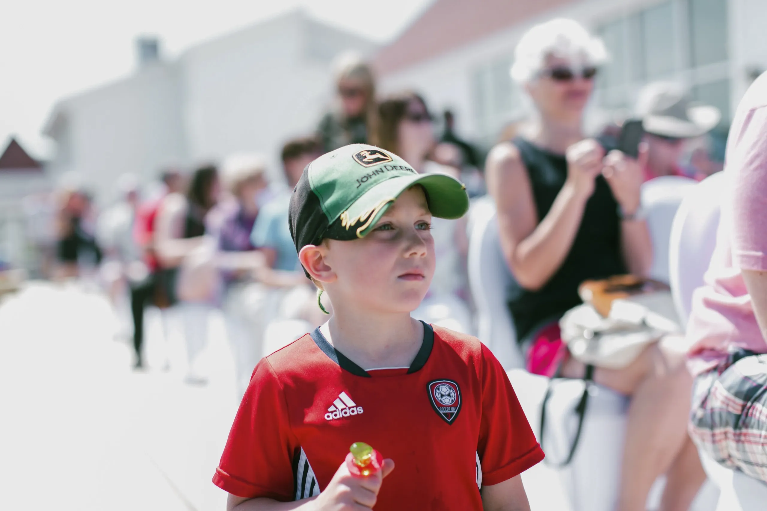 A young boy in a red soccer jersey and green baseball cap outdoors, with blurred guests and white buildings behind him.